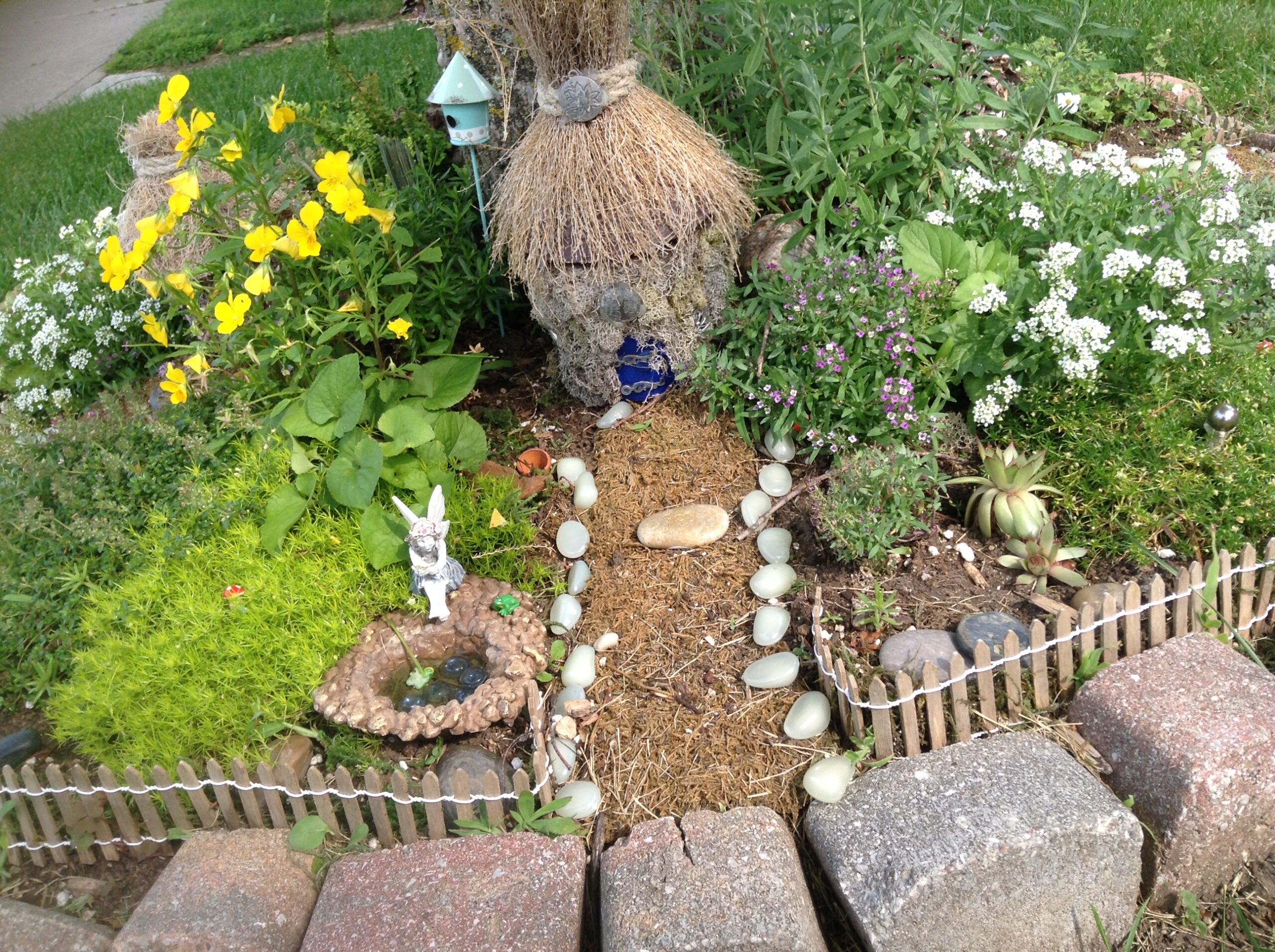 Miniature garden at the base of a tree. Figurines, pebbles and fence complete the garden.
